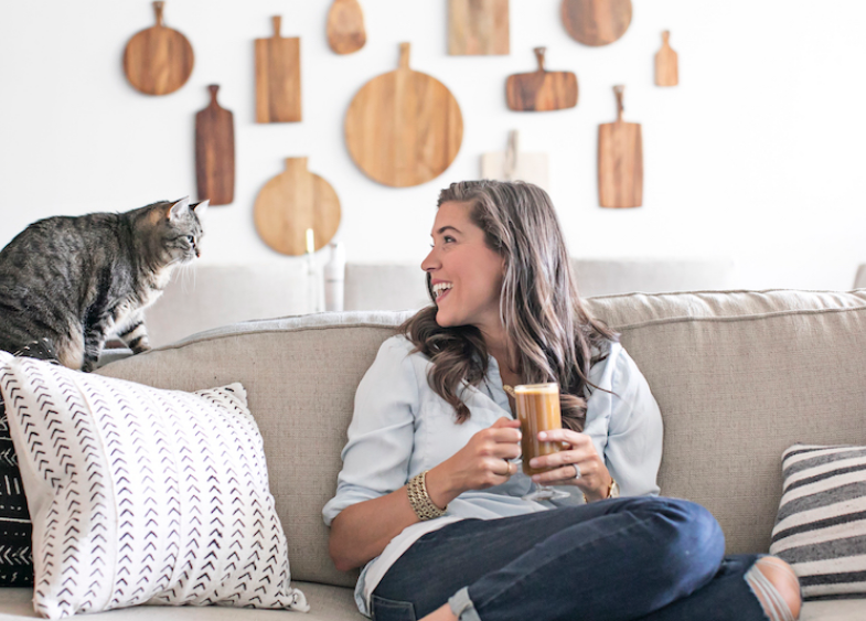 light-skinned woman sitting on a couch, holding coffee, looking at her cat