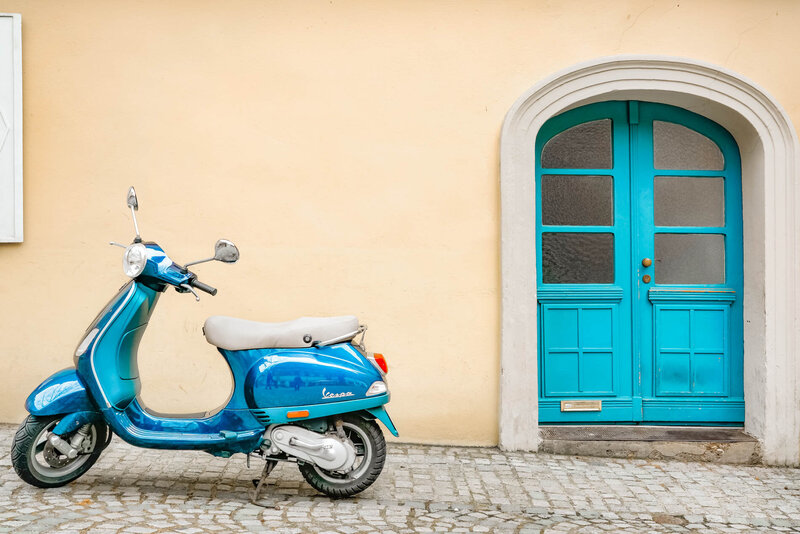 An Italian street with cobblestones featuring a yellow building with a blue door and in front of it sits a blue moped.