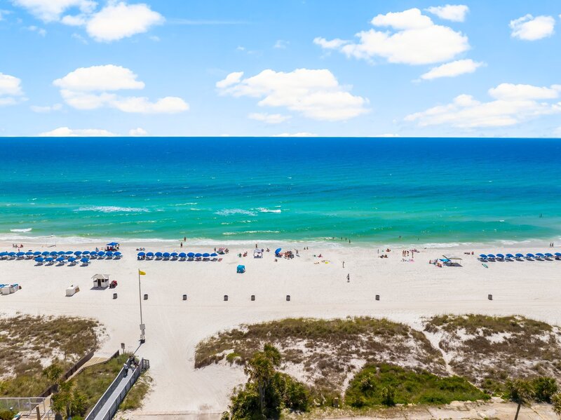 Aerial view of the white-sand beach and emerald-green waters directly across from The Surf House vacation rental in Panama City Beach, Florida, with rows of blue beach chairs and umbrellas lining the shoreline.