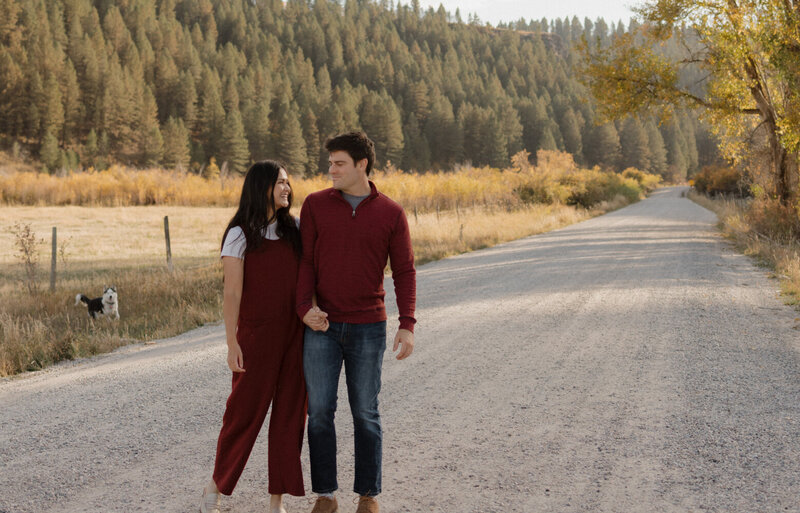 A husband and wife are holding hands and looking at each other smiling. They're outside, with lots of pine trees behind them.