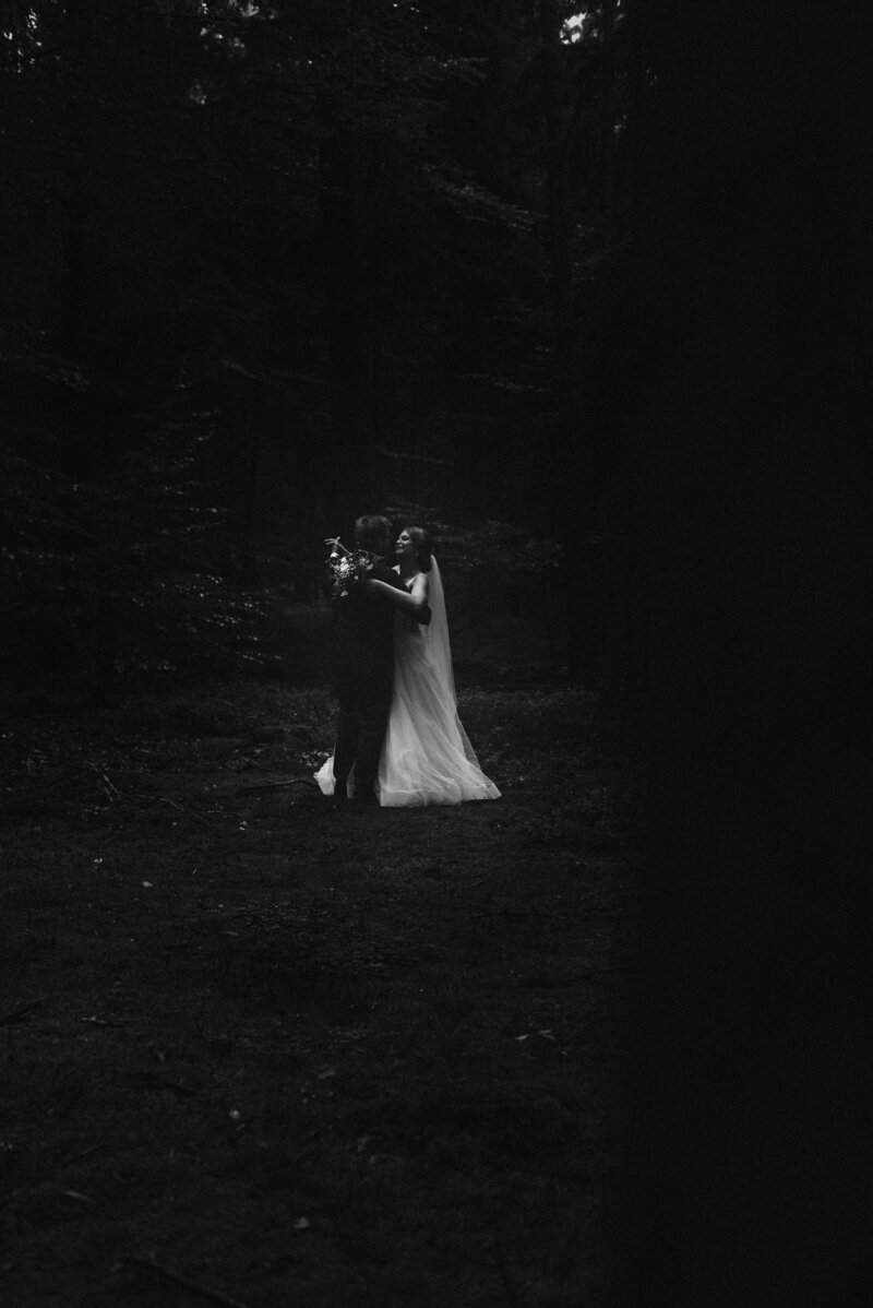 Dunkle schwarz-weiß Aufnahme eines Brautpaars im Wald, bei der das Brautkleid der einzig helle Punkt im Bild ist.