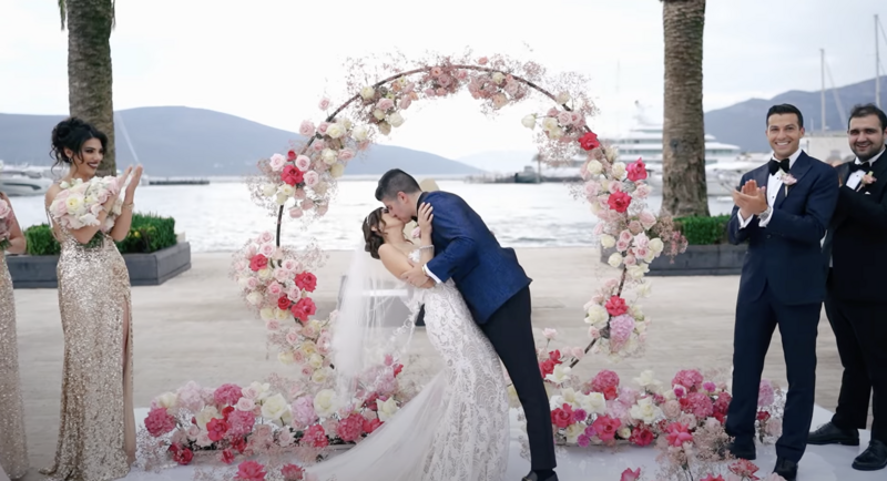 Groom kisses bride under a pink flower arch by the Adriatic Sea, with groomsmen applauding nearby — emotional moment from a luxury wedding film shot in Montenegro