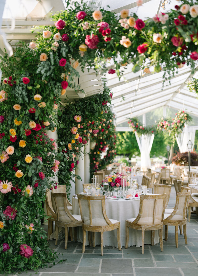 Outdoor wedding reception area decorated with lush rose arches in various colors, wooden chairs, and tables set with colorful floral centerpieces.