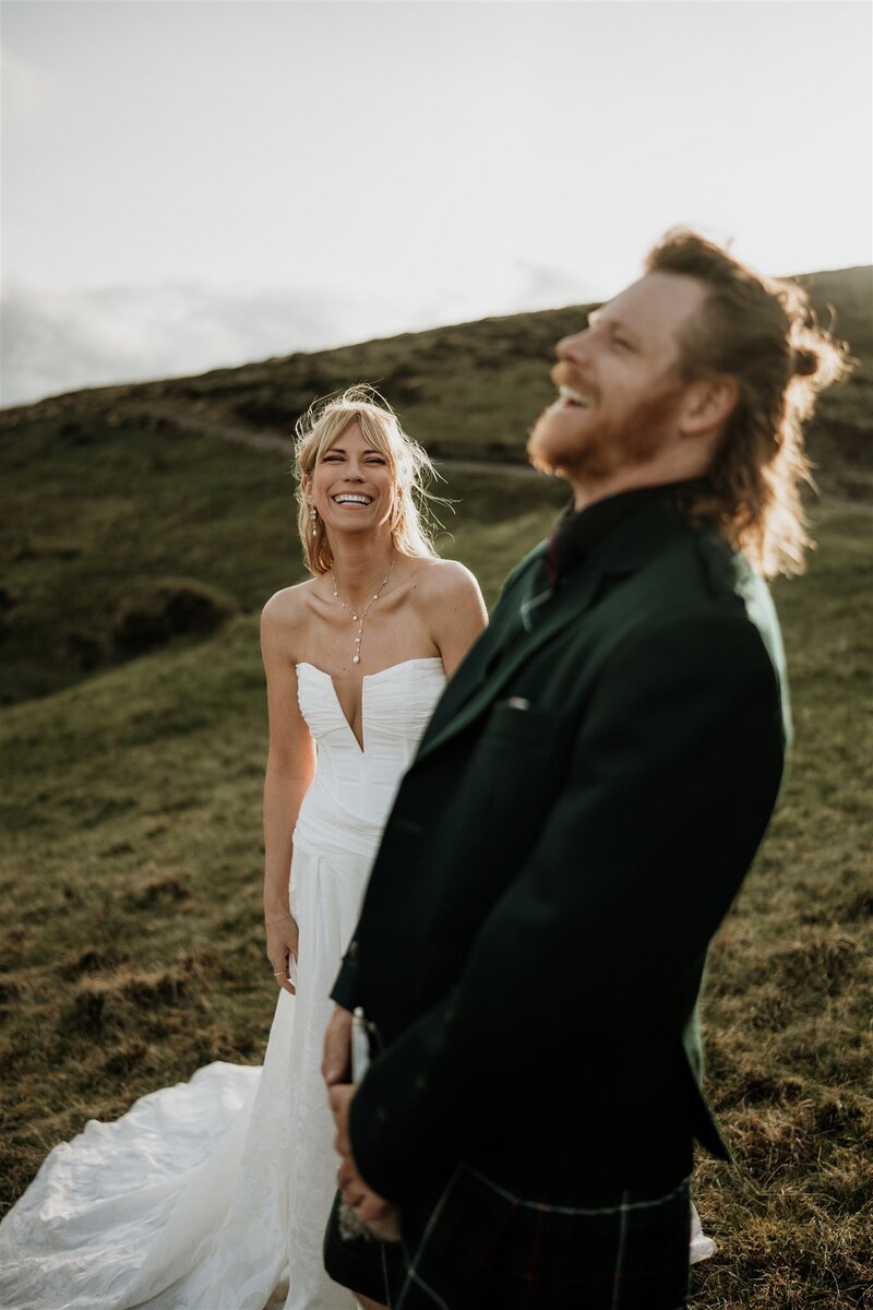 A bride and groom laugh candidly at each other as they’re photographed in a wide Aberdeen landscape. Green hills can be seen behind them as the sun starts the set. They both wear wedding attire - a dress and a kilt.