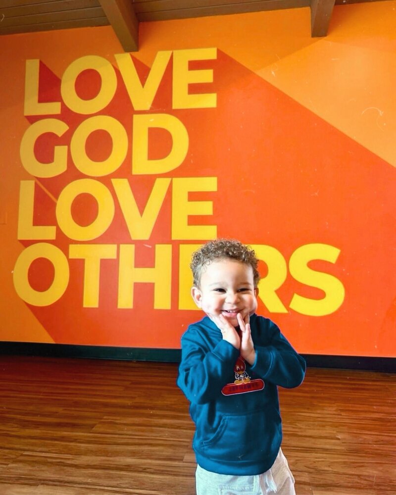 Little child smiles while standing in front of wall that says Love God Love Others