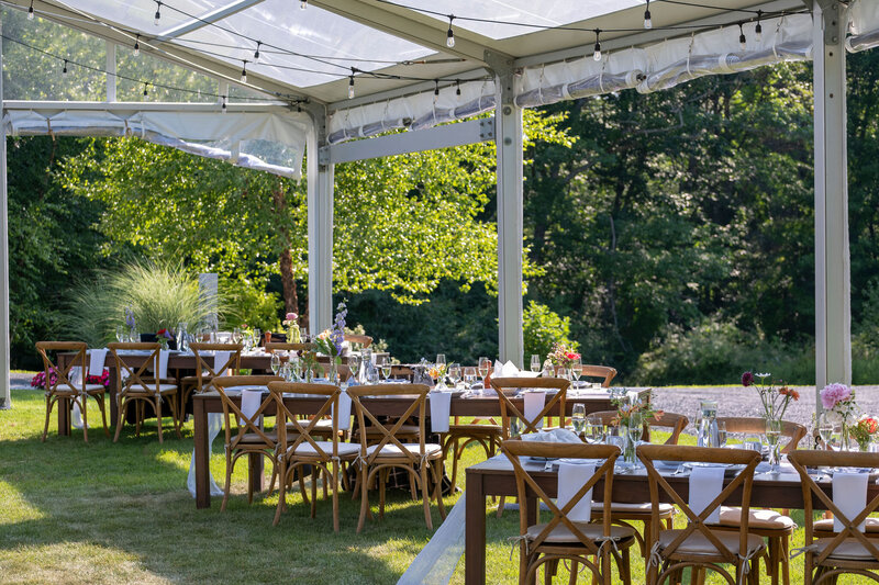 In a clear tent, sets 3 wooden farm tables, all set with white runners, glasses, different flowers, and plates. Wooden chairs sit on both sides of each with beige cushions