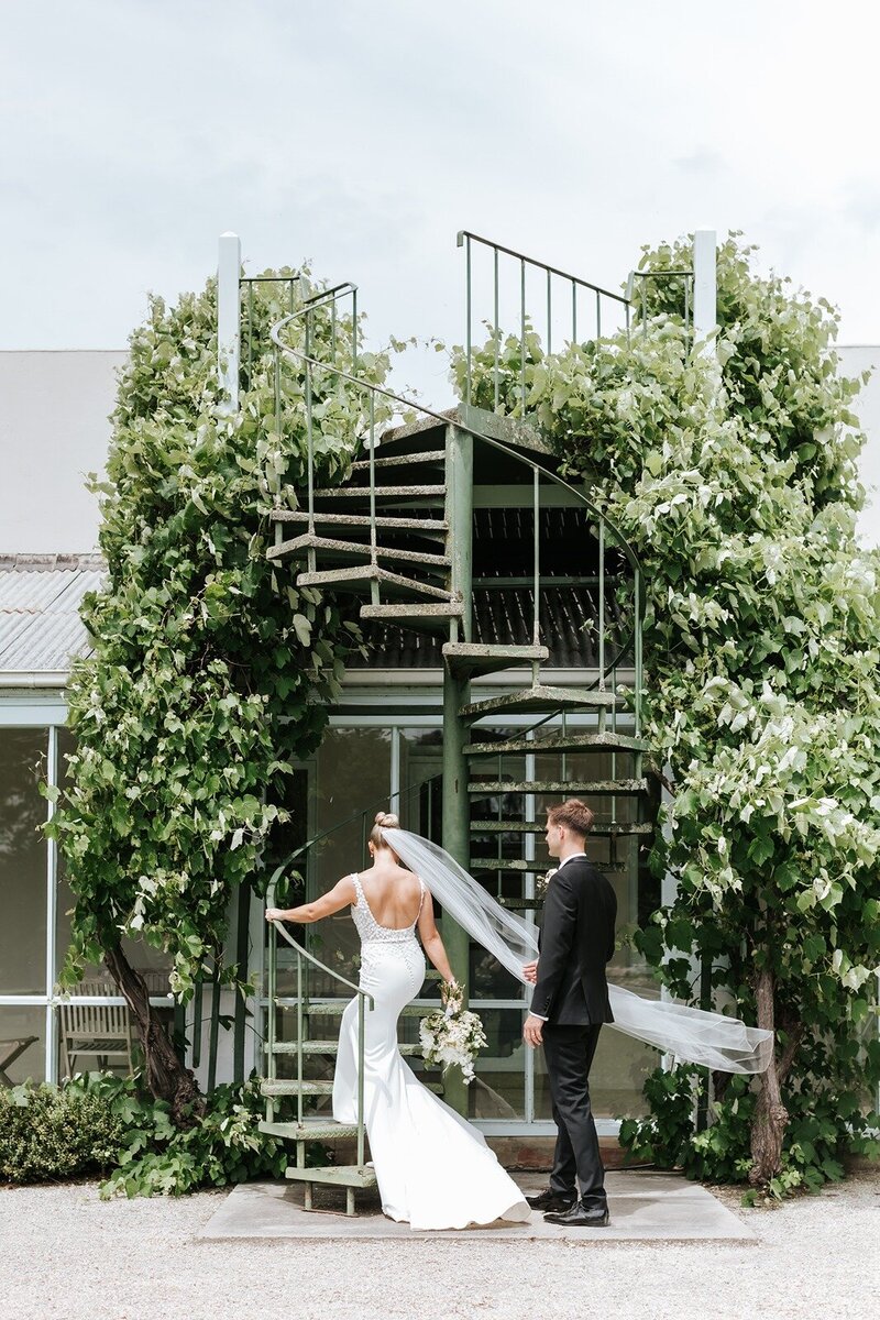 A bride with a white dress and long veil walking up a green spiral staircase with her groom.