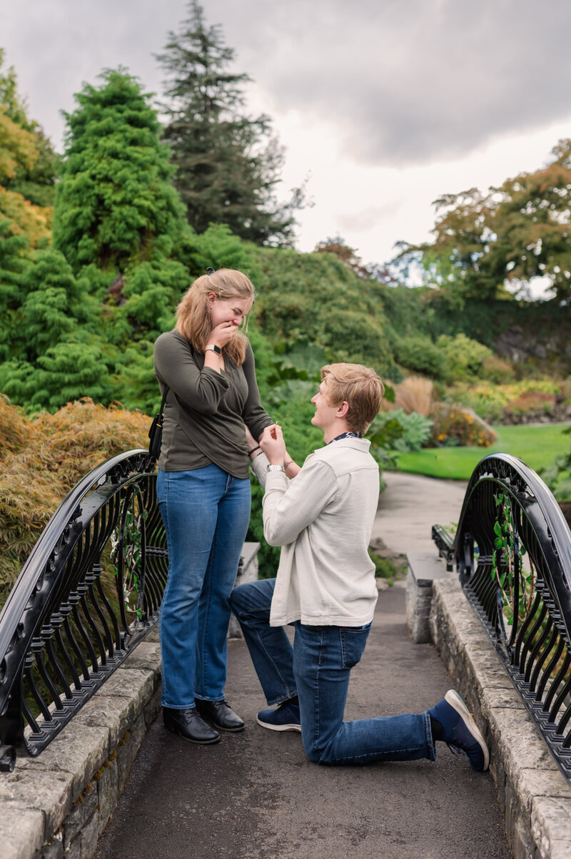 newly engaged couple kissing on a bridge at Queen Elizabeth Park. Girls left foot extended out while couple kiss.