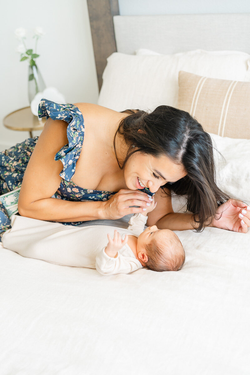 a mother lays on a bed and leans over her newborn baby while smiling and holding his hand while captured by a Leander photographer.