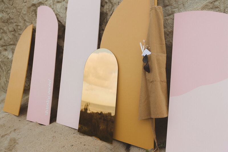 Custom-made backdrops by Shindig Social arranged against a rock formation. The backdrops feature various shapes and colors, including a reflective gold panel, pink, white, and beige panels. A beige apron with paintbrushes and sunglasses hangs on one of the backdrops.