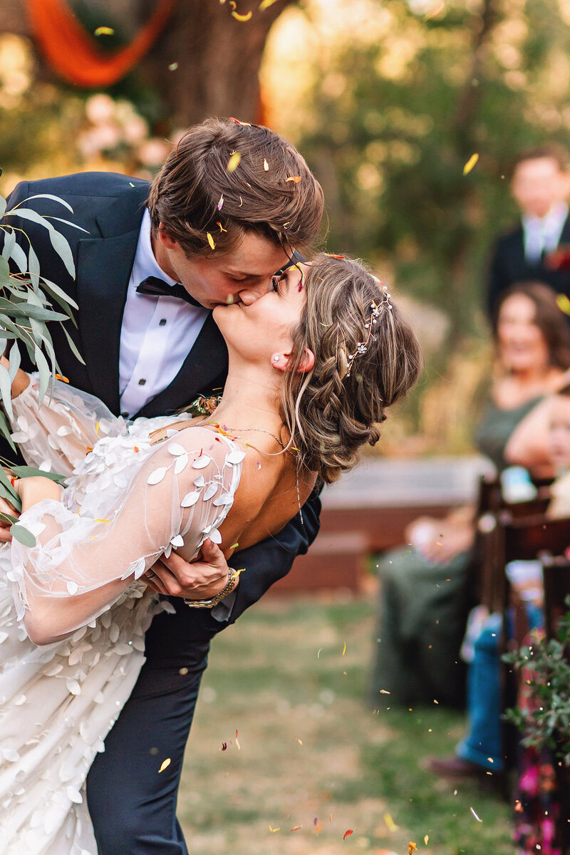 groom dips bridal while guests toss flower petal confetti during their walk up the aisle