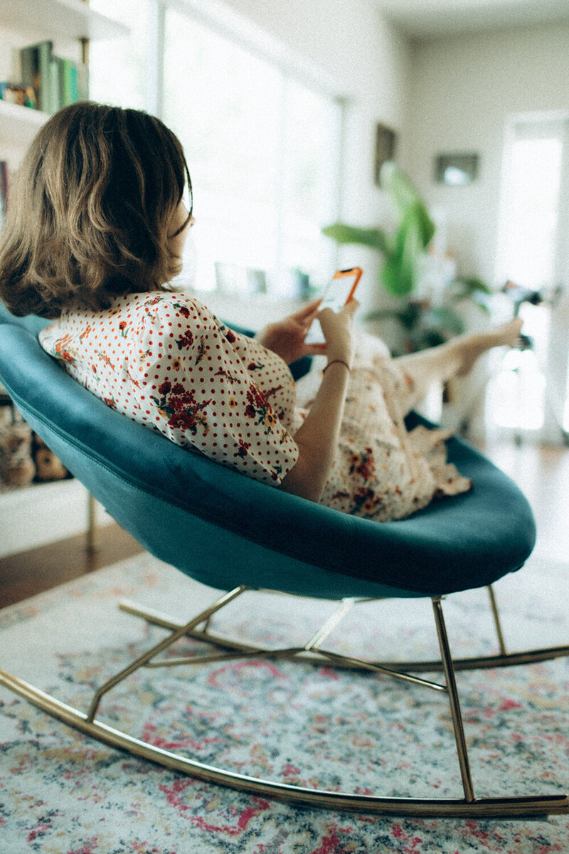 Andrea Shah sitting in a blue chair, relaxing and looking at her phone, wearing a floral dress
