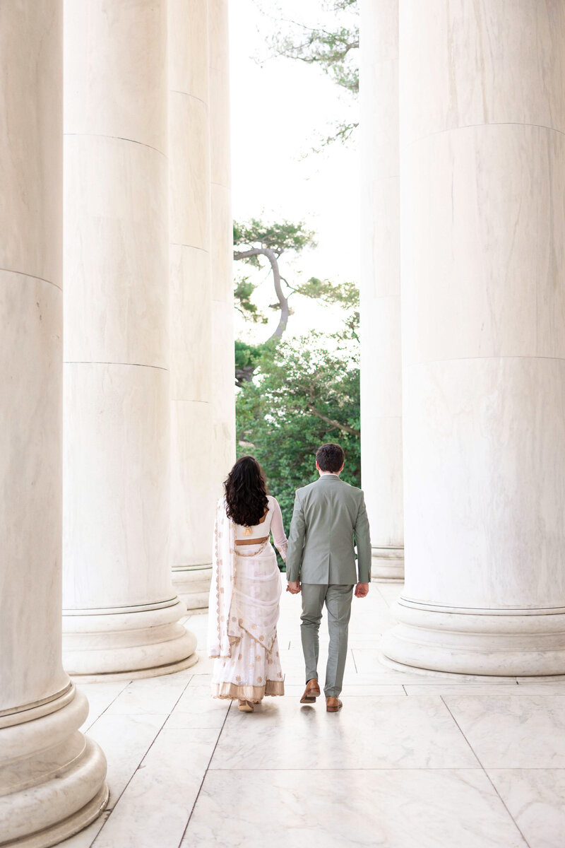 A couple walks hand in hand between tall marble columns. The woman is wearing a white and gold sari, and the man is in a light gray suit. They are surrounded by greenery and the scene is serene.