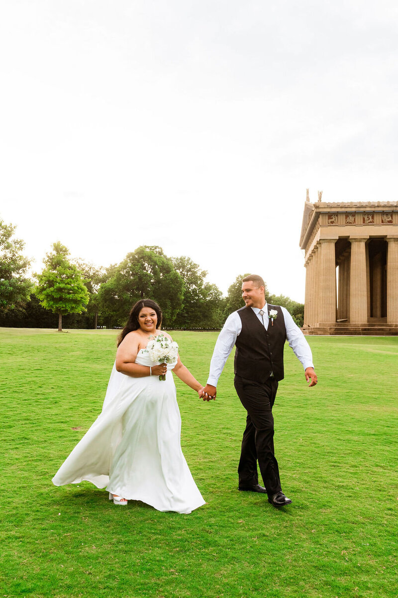 Groom smiling at his new wife who is holding her flowers and smiling at the camera as they walk outside the Nashville parthenon building