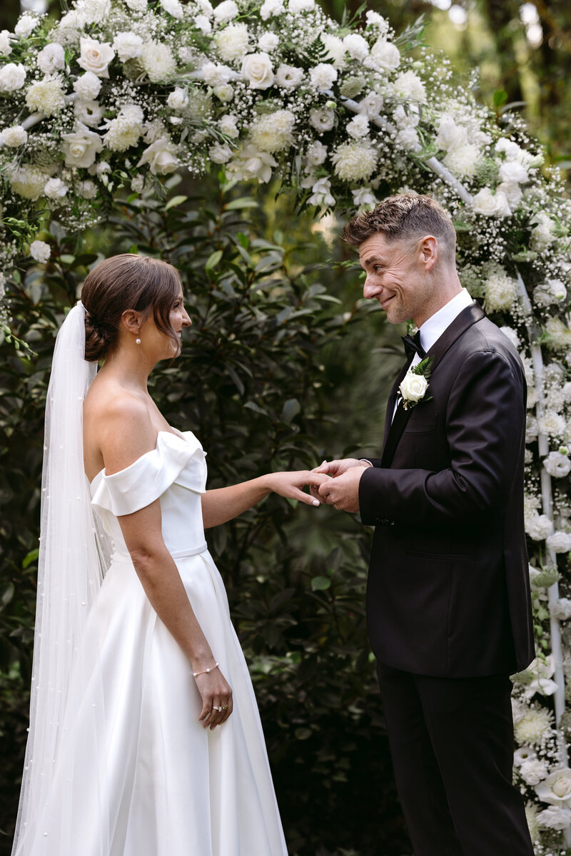 Bride and groom exchanging rings during their elegant wedding ceremony at Willow and Stone Estate, captured by Carry Your Heart Photography. The intimate moment is beautifully framed by the estate's charming garden setting, with lush greenery and blooming flowers enhancing the romantic atmosphere. This photo captures the couple's love and commitment as they say their vows in the picturesque surroundings of Willow and Stone Estate.