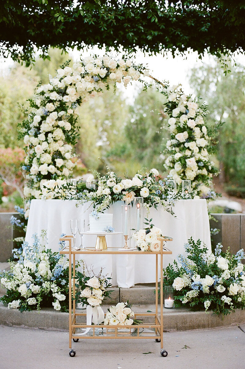16-radiant-love-event-outdoor-ceremony-floral-whiterose-arch-behind-sweetheart-table-romantic-greenery-white-roses-gold-bar-cart-cake-on-top-romantic-elegant-timeless