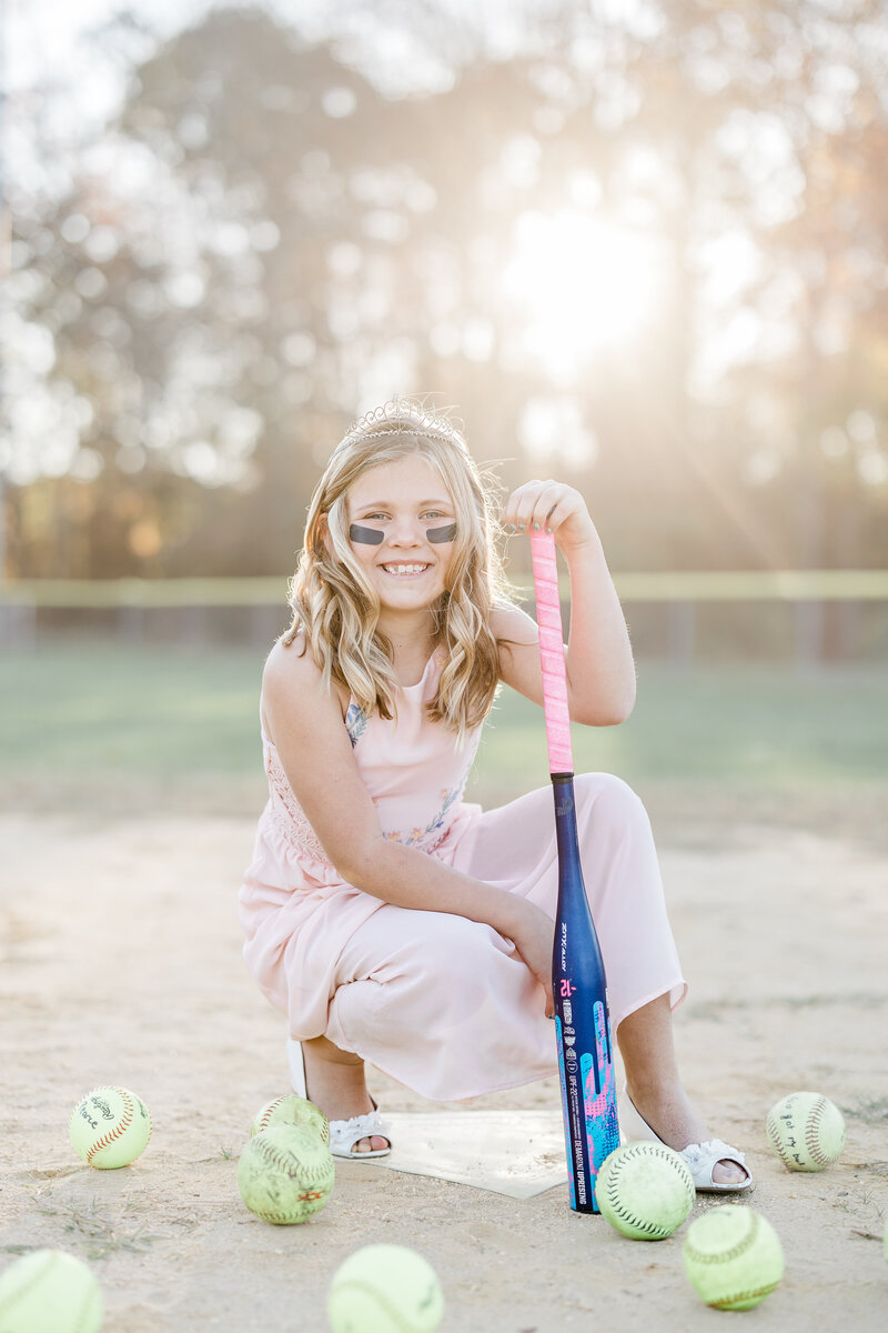 A young girl leans on a softball bat surrounded by balls at home plate at sunset