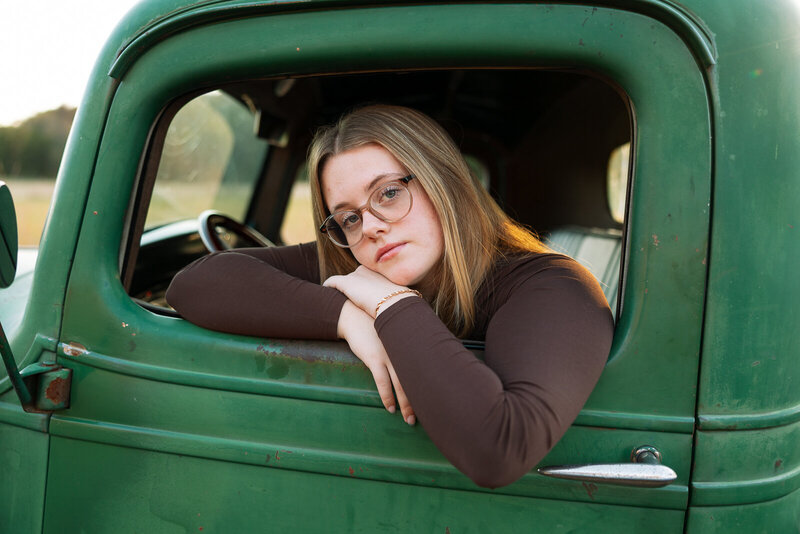 High school senior girl sitting in old green truck leaning in window looking at the camera in De Soto, Missouri.