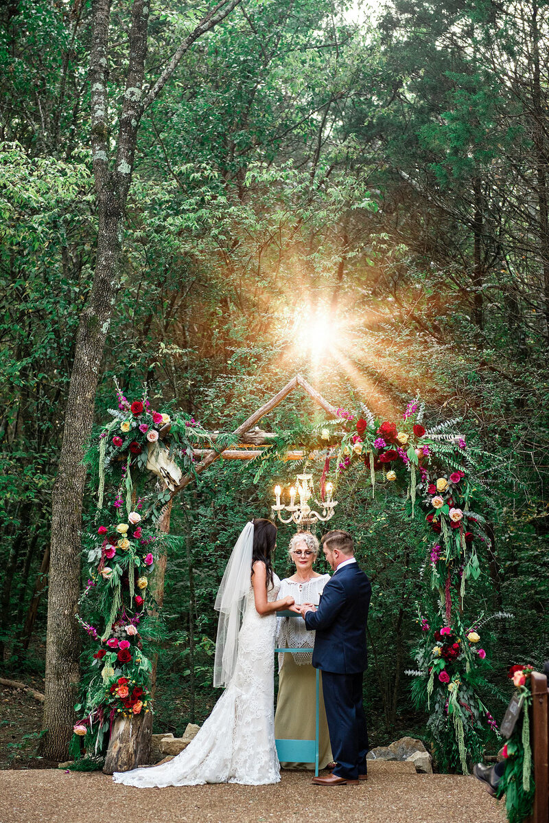 Couple exchanging rings at Drakewood Farm under arbor covered in bright colored flowers at sunset with sun shining through the trees