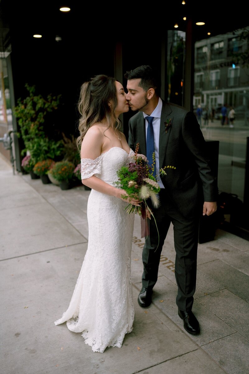 A couple in wedding attire sharing an affectionate kiss on a city sidewalk.