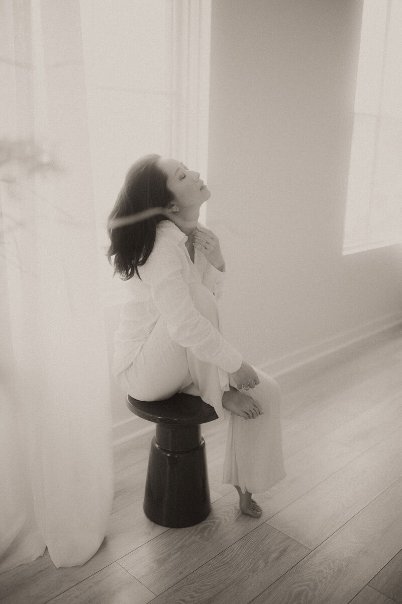 A woman in all white sitting on a stool in front of a window. 