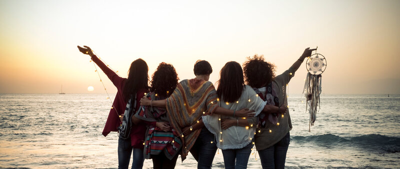 Back view of women holding each other at the water’s edge, evoking belonging, empowerment, and soulful collective energy