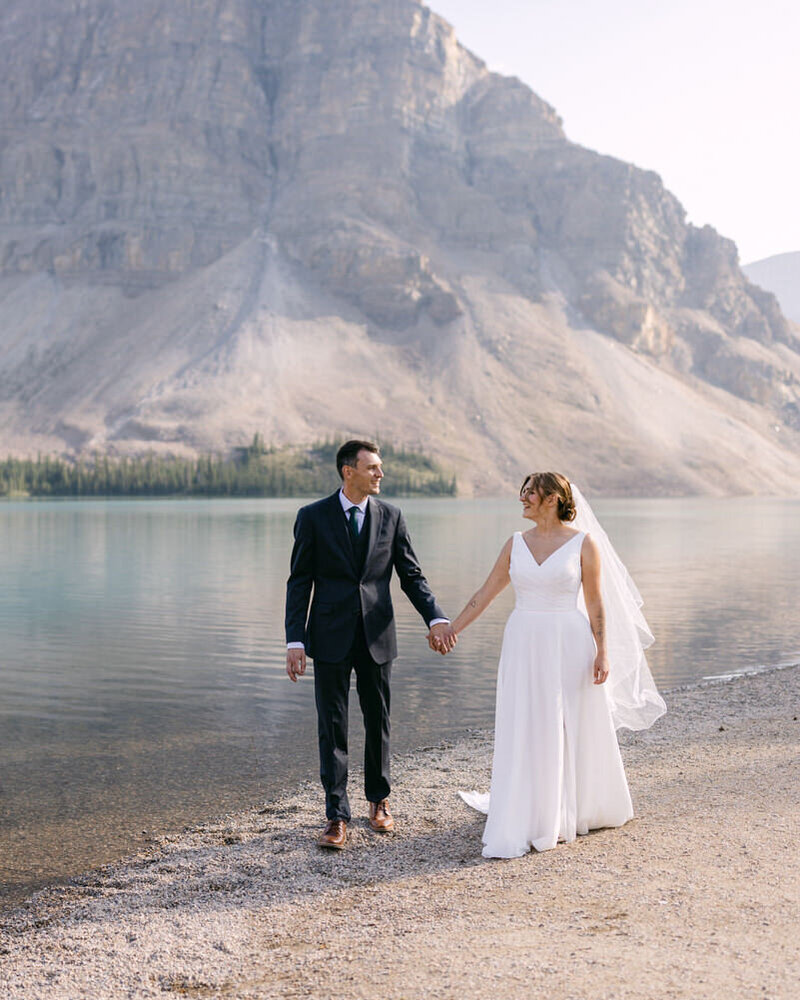 This romantic photograph captures the bride and groom walking hand in hand along the serene shoreline of Bow Lake in Banff National Park. The still waters and warm evening light create a stunning backdrop, adding a sense of peace and beauty to their intimate moment.