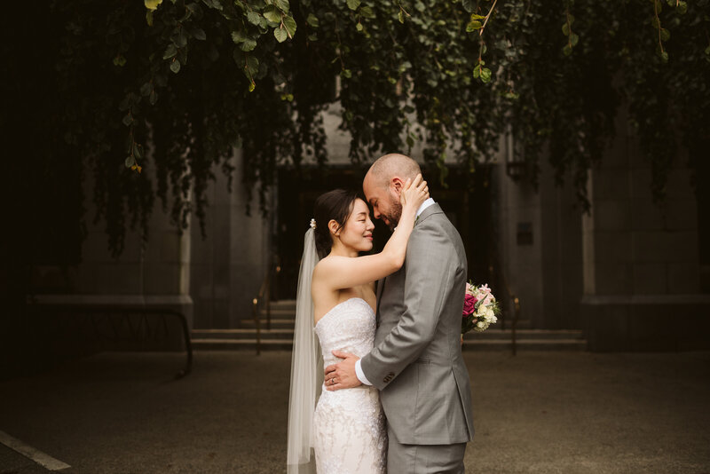 A couple embracing in front of Vancouver City Hall in their wedding attire.