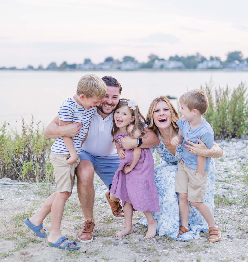 A family of five laughs and embraces on a beach at sunset during a summer photo session. The parents kneel down while their three young children surround them, full of energy and giggles. Captured by Nicole Neira Photography in Fairfield County, CT, this candid moment radiates joy, connection, and carefree coastal vibes.