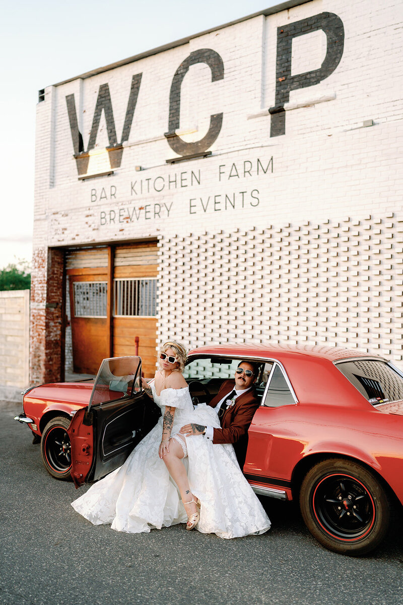 Vintage inspired Whitechapel Projects Wedding photo of a groom sitting in a vintage red car with the door open and the bride sitting on his lap. Taken in front of the white brick Whitechapel Projects Venue