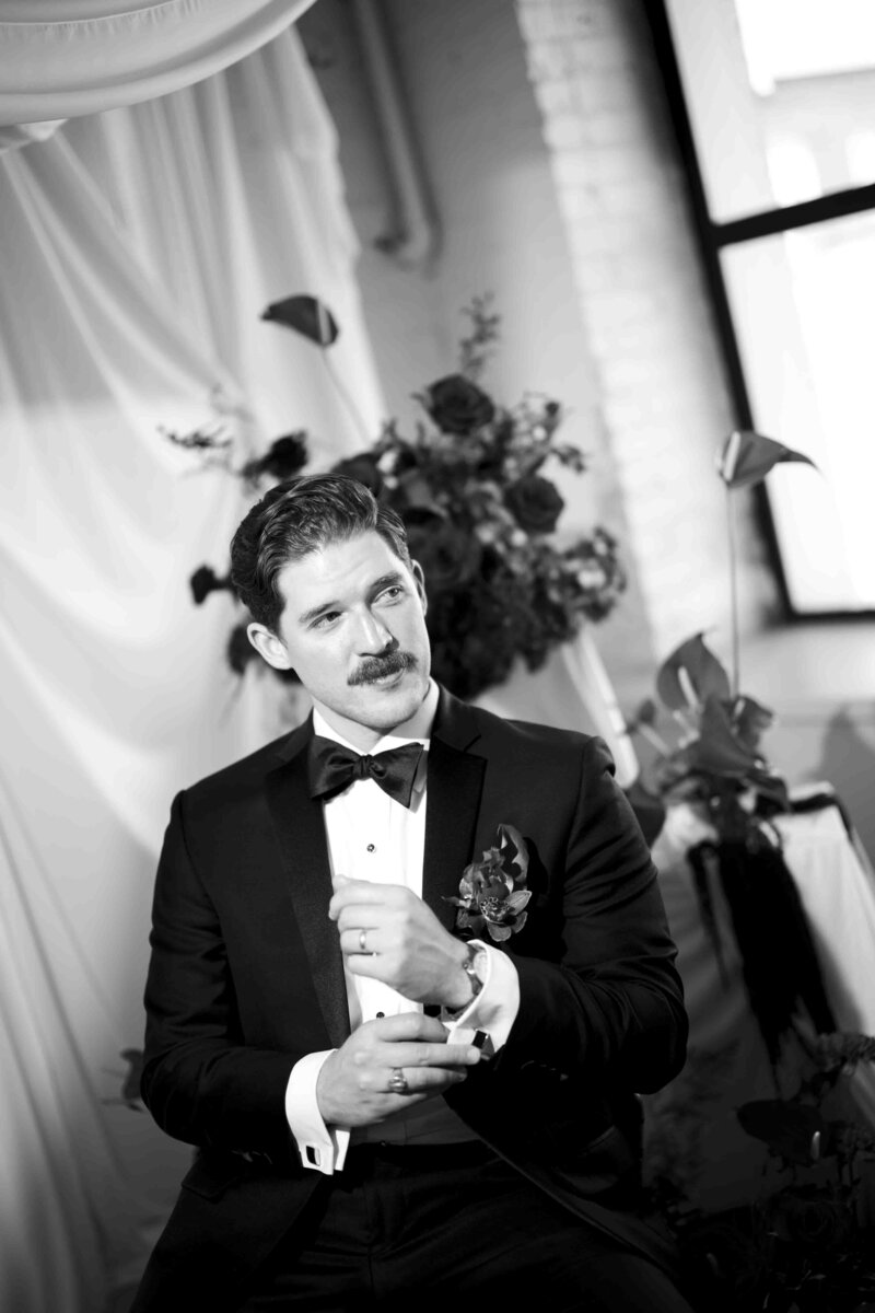 Editorial black and white photo of a groom adjusting his cuff links in front of a floral background. 