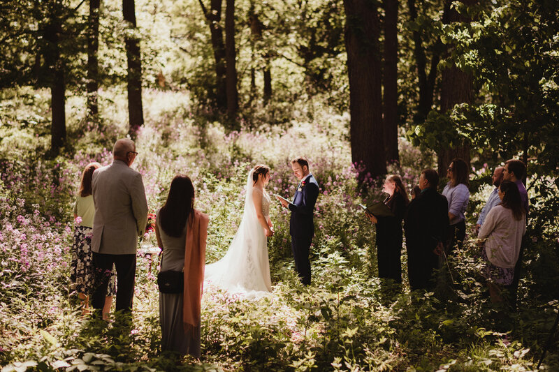 Bride and groom exchange vows during wedding ceremony in the woods as friends look on