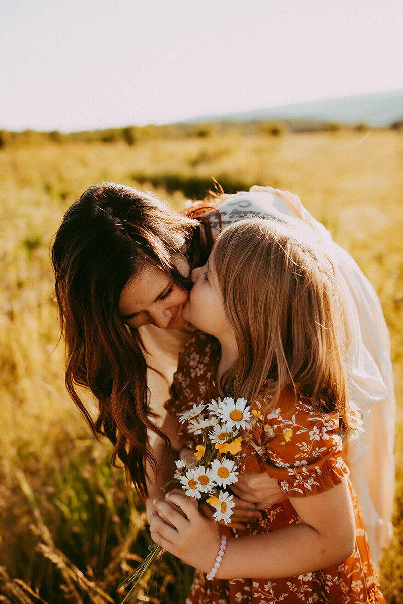 a daughter kissing her moms cheek as shes holding wildflowers standing in a field