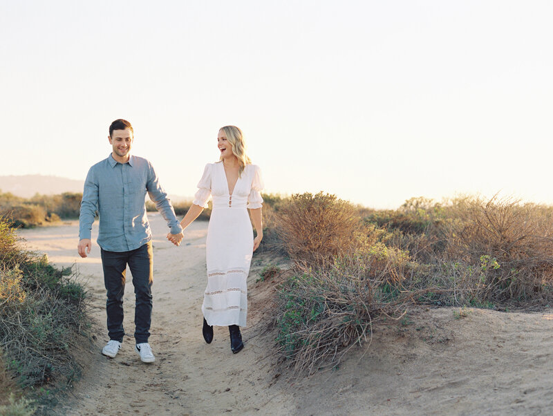 couple walking at the dirt path glider port in la jolla with white reformation midi dress and black boots