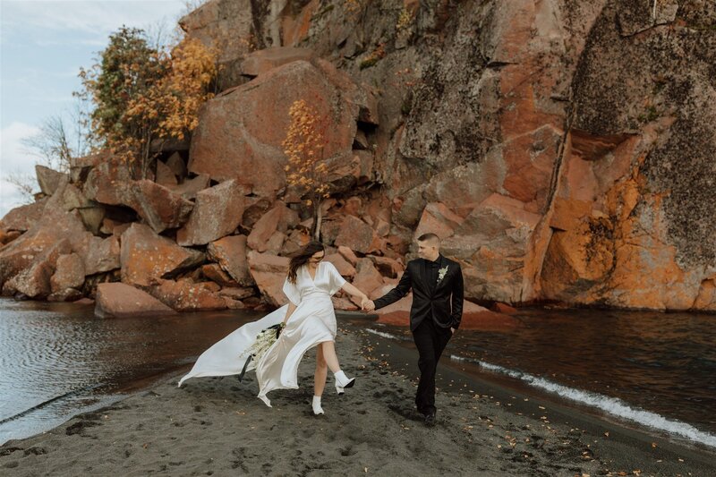 couple eloping on the north shore at black beach. bride and groom are holding hands and walking on the beach. bride is kicking up sand with her boot.