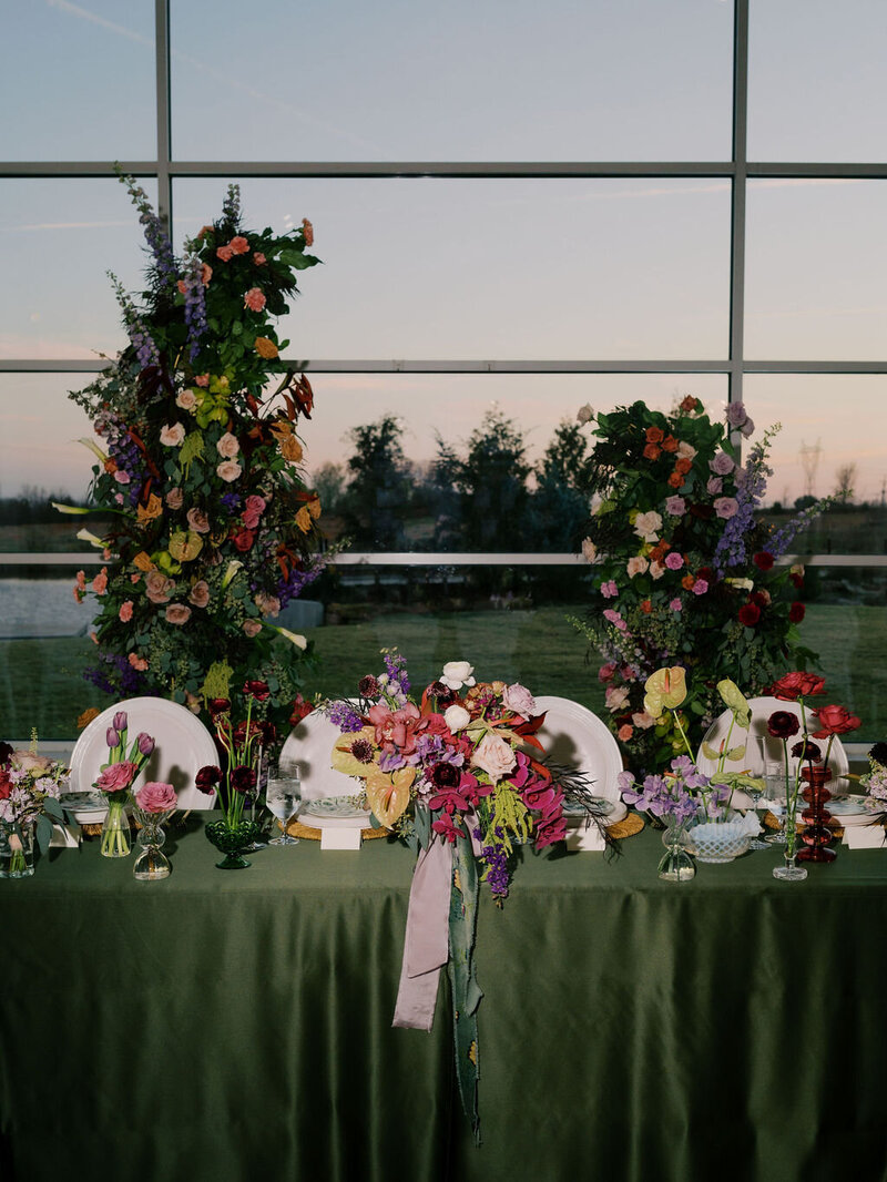 A direct flash photo of a colorful wedding reception tablescape, with green linens, and lush, eclectic florals of pink, magenta, purple, and green.