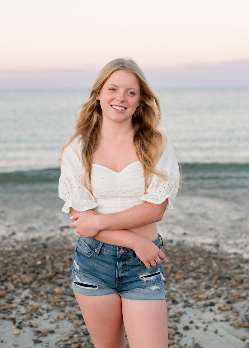 girl standing on the beach in front of the water with blonde hair, wearing a white shirt and jeans