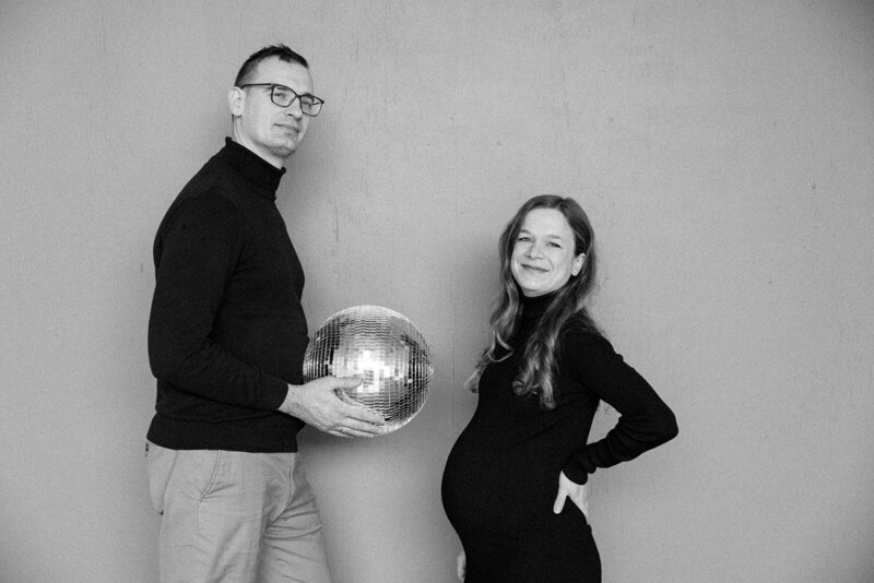 Man holds disco ball as woman holds pregnant belly in studio photo session. 