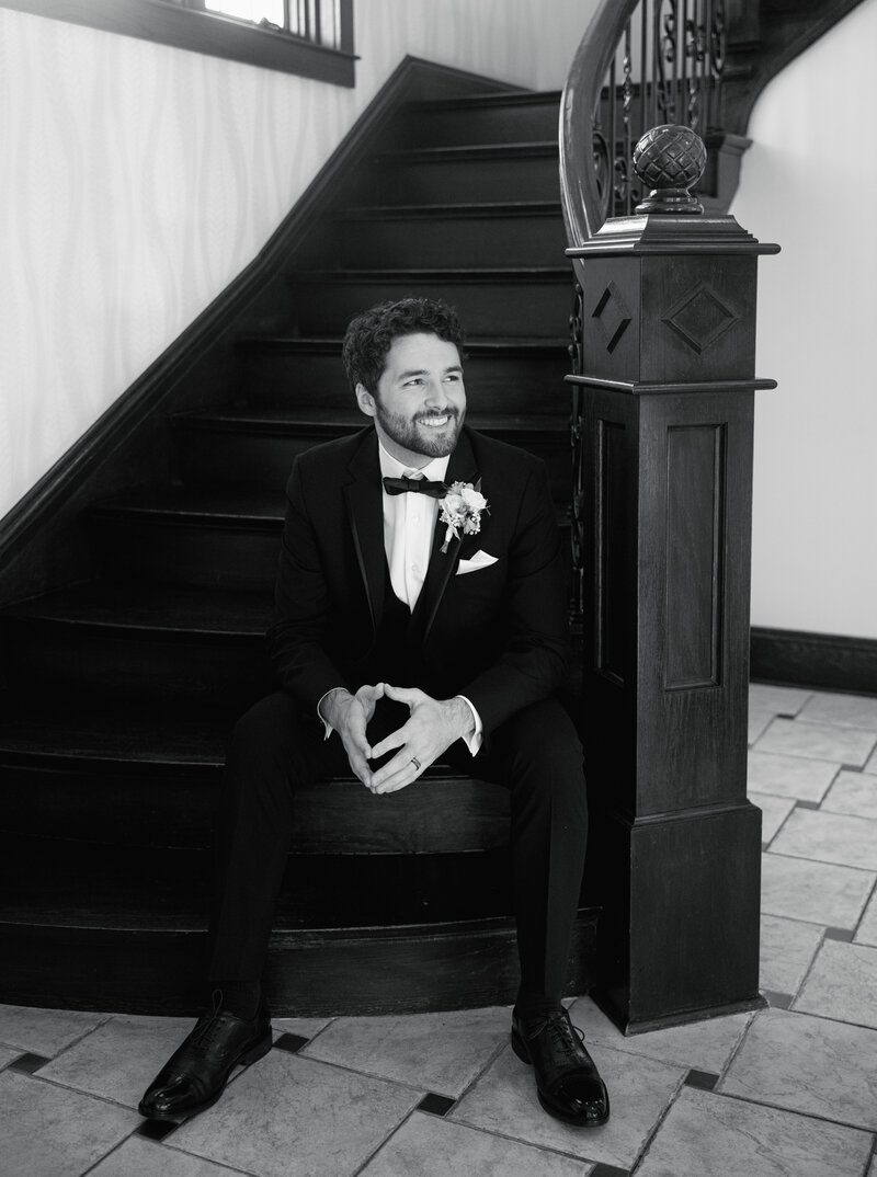 A black and white portrait of a groom sitting on a staircase in a tuxedo with black bow tie
