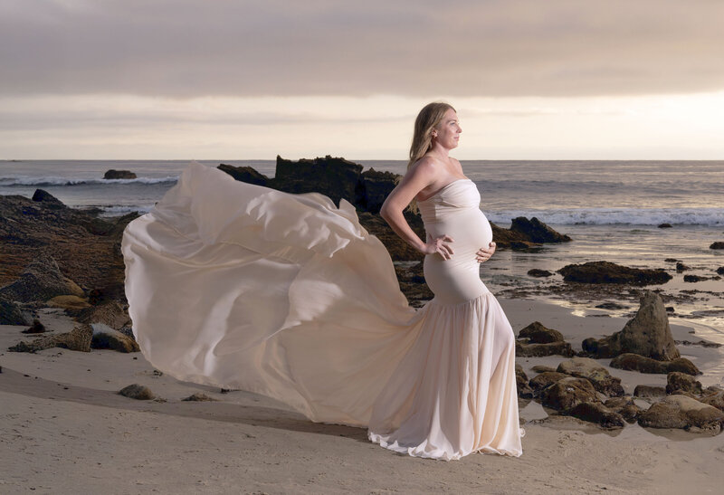 A mom to be stands on a beach with her pink maternity gown train flowing in the wind behind her