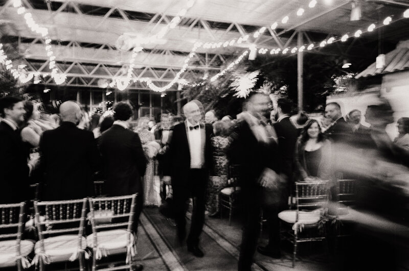 Black and white film photo with intentional motion blur showing guests mingling beneath string lights at a wedding reception at Roger Williams Park Botanical Center in Rhode Island — made by a film wedding photographer.