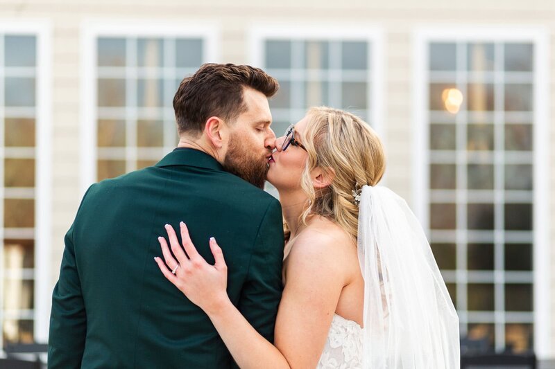 The bride, her veil cascading down her back, leans into her groom for a kiss, glasses slipping slightly as they press close. The groom’s dark green suit contrasts against the soft glow of the venue’s large-paned windows, capturing the quiet intimacy of their wedding day