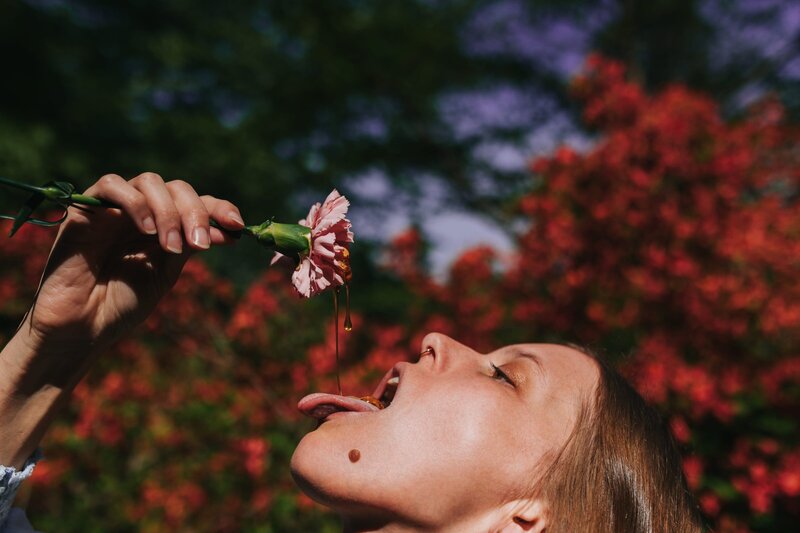 Woman drips honey from pink flower into her open mouth in front of tree with vibrant burgundy flowers.