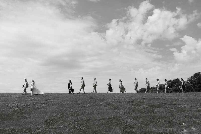 Black and white photo of a wedding party walking across an open field beneath a dramatic sky, captured in cinematic documentary style by Rainstorm Photo & Video.