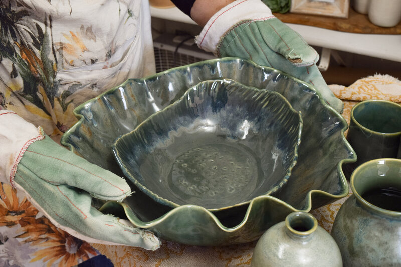Jill uses gloves to take two green serving dishes out of her kiln to sit beside handmade oil bottles and vases