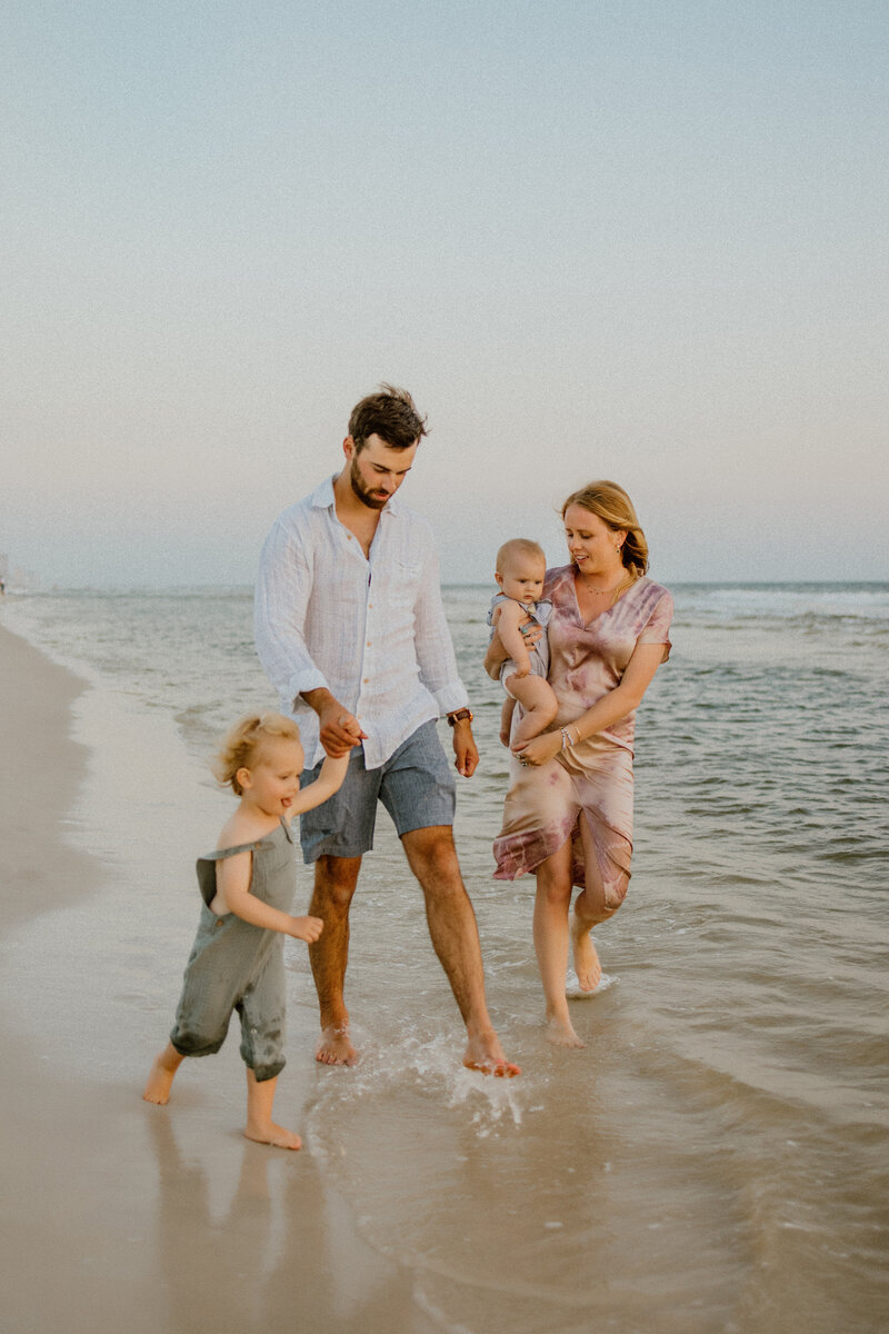 Family Photographer,  a you ng mother and father walk with their children, mom holds baby, and dad holds daughter's hand at the beach