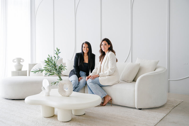 Courtney and Valerie, co-founders of Interiors by CV, sitting together in a modern living room with neutral furniture and minimalist décor.