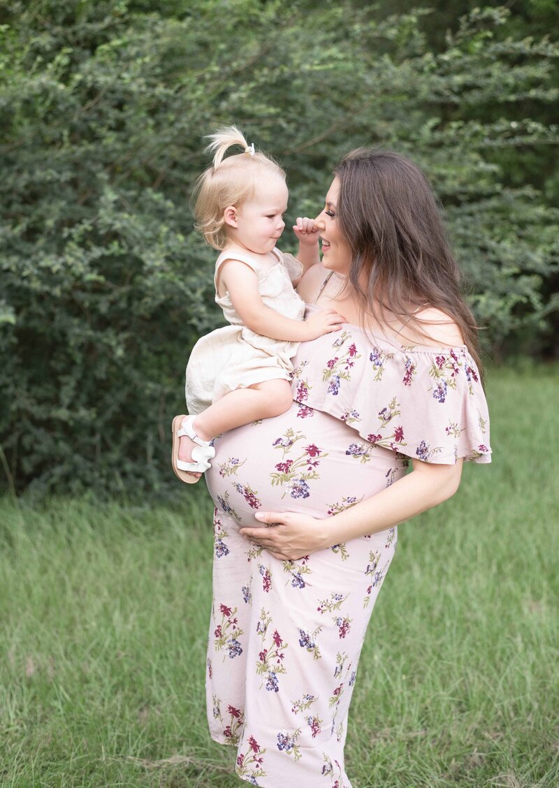 Expecting mother in a floral dress lifts her toddler in a grassy field, sharing a joyful moment. A maternity session by Moments in Light Photography.