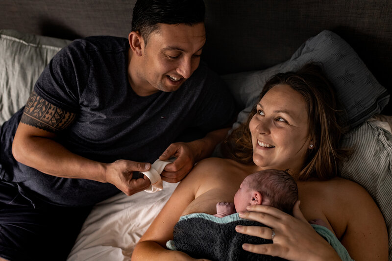 Een mooie thuisbevalling, moeder ligt met haar pasgeboren zoon op de borst en vader en moeder kijken elkaar verliefd aan.