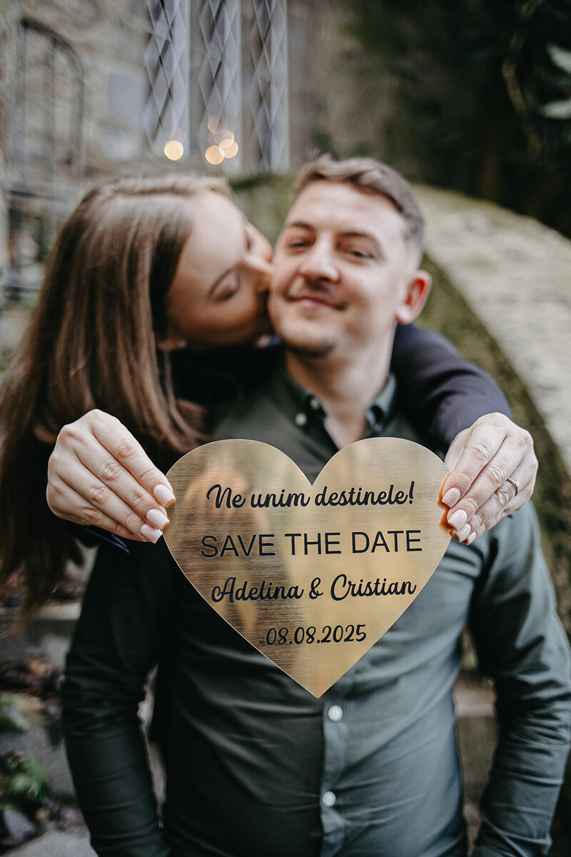 Bride-to-be kissing her fiancé on the cheek  in Malahide Castle, while holding a heart-shaped sign that says 'Save the Date' for their upcoming wedding